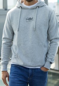 Homme portant un sweat à capuche gris clair avec poche avant et un jean bleu, debout à l'extérieur avec une main dans la poche.