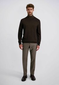 Man staat in een donkerbruin turtleneck sweater, grijze broek en donkerbruine leren schoenen tegen een witte achtergrond.