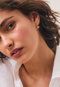 Gold heart-shaped earrings with a polished finish, hanging from a thin hoop. Model wearing a white collared shirt, slight side profile.