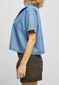 Woman wearing a loose light blue short-sleeve denim shirt and dark brown high-waisted denim skirt, shown from the side against a plain background.