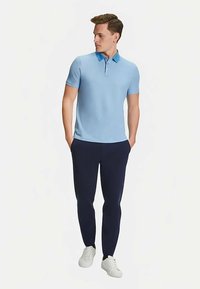 Lichtblauw poloshirt met korte mouwen en een kraag, gecombineerd met marineblauwe broek en witte sneakers, staand met handen in de zakken.