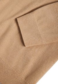 Hihapuserin beige neuleen hiha ribbireunuksella, joka näyttää tekstuuria ja hienoa ompelua.