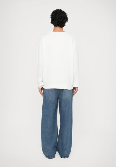 Sudadera blanca de corte relajado, con cuello redondo y puños de canalé, combinada con un pantalón vaquero azul holgado que cuenta con bolsillos traseros y una cintura alta.