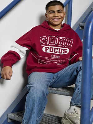 Bordo kapuce ar baltajiem akcentiem un grafisko tekstu, kombinēta ar gaiši ziliem džinsiem. Sēž uz metāla kāpnēm, smaidot, ar īsiem matiem un baltām sporta apaviem.