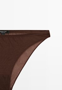 Brun bomulds-thong med en glat tekstur, der har et minimalistisk design og en talje-elastik. Mærkeetiketten er synlig på venstre side.