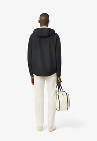 Giacca nera con cappuccio, dal texture morbido, indossata sopra pantaloni bianchi. Porta una borsa color crema con dettagli neri e cerniera.