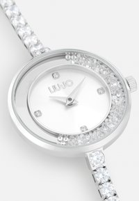 Reloj de pulsera plateado con caja redonda, esfera blanca, cristales alrededor del bisel y una pulsera con acentos de cristal. Presenta marcadores de hora minimalistas.