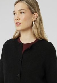 Cardigan noir en tricot avec un col rond, tissu texturé et cinq boutons. Porté sur un haut bordeaux, avec des boucles d'oreilles en goutte de perle.