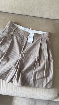 Beige pinstriped shorts with pleats, featuring a belt loop waistband and a white lining. Tag attached with care instructions.