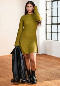 Olivengrünes langärmeliges Kleid aus strukturiertem Stoff mit einem seitlichen Schlitz, kombiniert mit einer schwarzen Lederjacke und klobigen schwarzen Stiefeln.