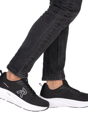 Osoba nosząca czarne siateczkowe trampki z białymi podeszwami i ciemnoszare dżinsy, idąca z lekko uniesioną jedną stopą.