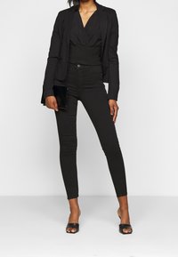 Blazer noir porté sur un haut noir texturé et un jean noir ajusté. Pochette noire à la main. Mules noires à bout ouvert.