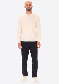 Uomo in piedi che indossa un maglione crema a maniche lunghe, pantaloni neri e sneakers bianche su uno sfondo chiaro uniforme.