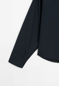 Manche longue d'une chemise à petits carreaux bleu foncé et noir avec poignet et ourlet courbé sur fond blanc.