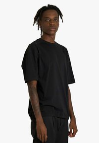 Giovane con dreadlocks che indossa una maglietta nera larga e pantaloni neri, mostrando un tatuaggio sull'avambraccio destro.