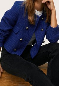 Koninklijk blauwe tweedjas met een cropped ontwerp, dubbelbrede gouden knopen en twee voorzakken. Draagt over een wit shirt.