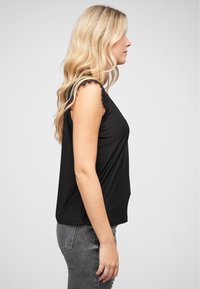 Top sans manches noir en tissu doux, avec des bordures en dentelle sur les épaules, associé à un jean gris ajusté.