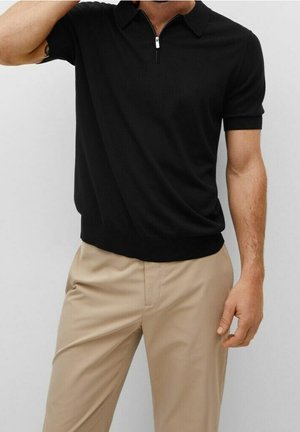 Polo noir à manches courtes avec un col zippé, fabriqué dans un tissu lisse. Associé à un pantalon beige clair.