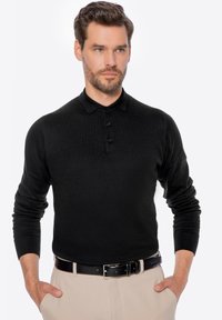 Schwarzes Langarm-Polohemd mit Kragen, ausgestattet mit einer Knopfleiste und taillierten Manschetten. Getragen mit hellen Hosen und einem schwarzen Gürtel.
