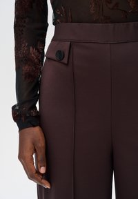 Close-up van een persoon in donkerbruine high-waisted broek met een zwart knoopdetail en een doorschijnende zwarte top met bloemborduursel.