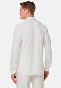 Camicia bianca a maniche lunghe con una texture liscia, colletto classico, cucitura del yoke posteriore e polsini con bottone, mostrata da un angolo laterale.
