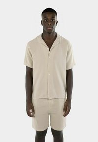 Lyse beige kortermet skjorte og shorts-sett, laget av strukturert stoff. Har knappefront og avslappet passform. Enkelt design uten mønstre.