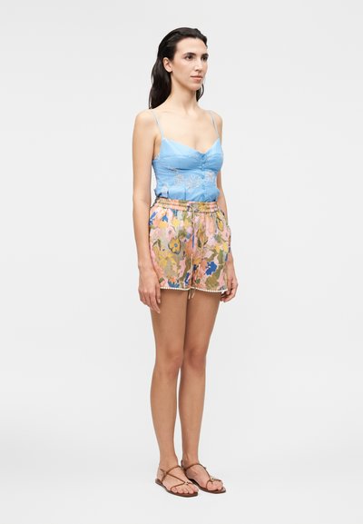 Γυναίκα που φοράει μπλε κεντητό τοπάκι, πολύχρωμα φλοράλ σορτς με κορδόνι και καφέ σανδάλια, στέκεται μπροστά σε ένα απλό ανοιχτό φόντο.