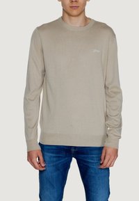 Maglione beige lavorato a maglia con collo rotondo, polsini e orlo a coste. Dettaglio del logo in modo sottile sul petto. Indossato con jeans blu.