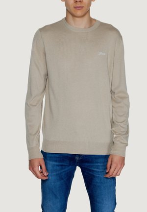 Maglione beige lavorato a maglia con collo rotondo, polsini e orlo a coste. Dettaglio del logo in modo sottile sul petto. Indossato con jeans blu.