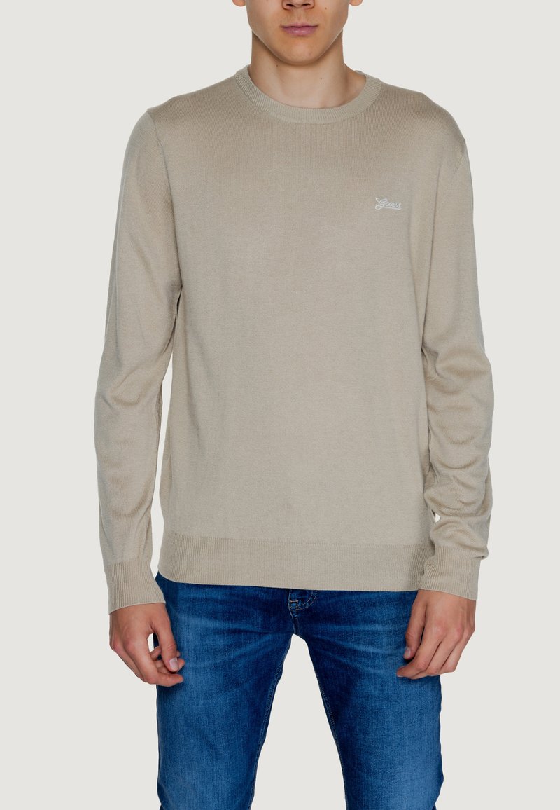 Maglione beige lavorato a maglia con collo rotondo, polsini e orlo a coste. Dettaglio del logo in modo sottile sul petto. Indossato con jeans blu.