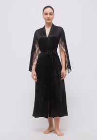 Γυναίκα που φοράει μαύρη σατέν ρόμπα με μανίκια κάπα διακοσμημένα με δαντέλα και στη μέση ζώνη δεμένη, στέκεται ξυπόλητη μπροστά σε μια απλή φόντο.