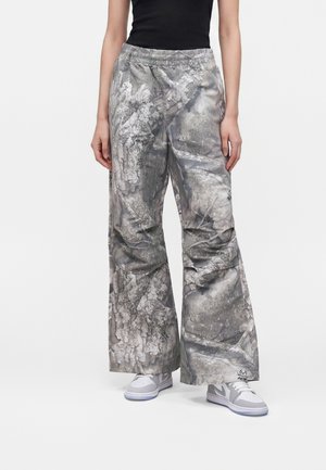 Persona che indossa pantaloni a gamba larga con motivo grigio strutturato e sneakers bianche su uno sfondo bianco uniforme.