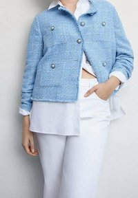 Veste en tweed bleu clair texturée avec des boutons argentés, dotée de deux poches avant, portée sur une chemise blanche et assortie à un pantalon blanc.