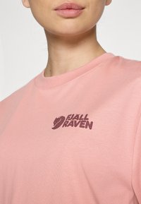 Camiseta de algodão rosa clara com decote redondo e um logotipo "Fjällräven" em rosa escuro na área do peito esquerdo. Textura suave.