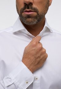 Witte overhemd met een spread collar, gladde textuur en knopenmanchetten. Zichtbare hand die de kraag aanpast.