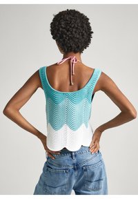 Person med lockigt hår som bär en ärmlös blå och vit stickad topp med vågiga kanter och lösa blå jeans, med händerna på höfterna, ryggen vänd mot kameran.