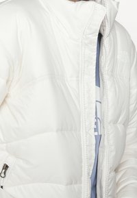 White insulated puffer jacket with a high collar, visible stitching, front zipper, and logo on the upper left side. Soft texture.