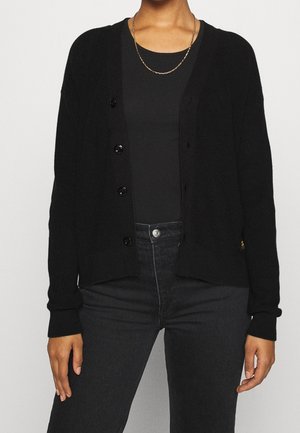 Zwarte gebreide cardigan met een knoopsluiting aan de voorkant, een geribbelde textuur, V-hals en lange mouwen, gecombineerd met een zwarte top en jeans.