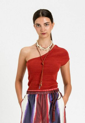 Giovane donna con capelli scuri indossa un top rosso monospalla e pantaloni tie-dye multicolori, con una collana di conchiglie e una collana di pelle.