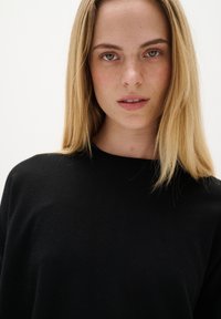 Schwarzer Pullover mit Rundhalsausschnitt. Weiche Textur, leicht lockere Passform und minimale Details. Model mit glattem, blonden Haar und neutralem Make-up.