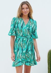 Wickelkleid mit einem lebhaften grünen und blauen Blättermuster, kurzen ausgestellten Ärmeln, einem Bindegürtel und einem Rüschensaum. Leichter Stoff.