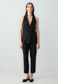 Completo in lino nero caratterizzato da un blazer senza maniche con scollatura a V profonda e un solo bottone, abbinato a pantaloni a sigaretta e scarpe nere basse.