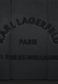Fekete textúrájú bőr, amelyen dombornyomott szöveg ível "Karl Lagerfeld" felirat alatt "Párizs" szerepel, alatta pedig félig látható "21 Rue St-Guillaume".