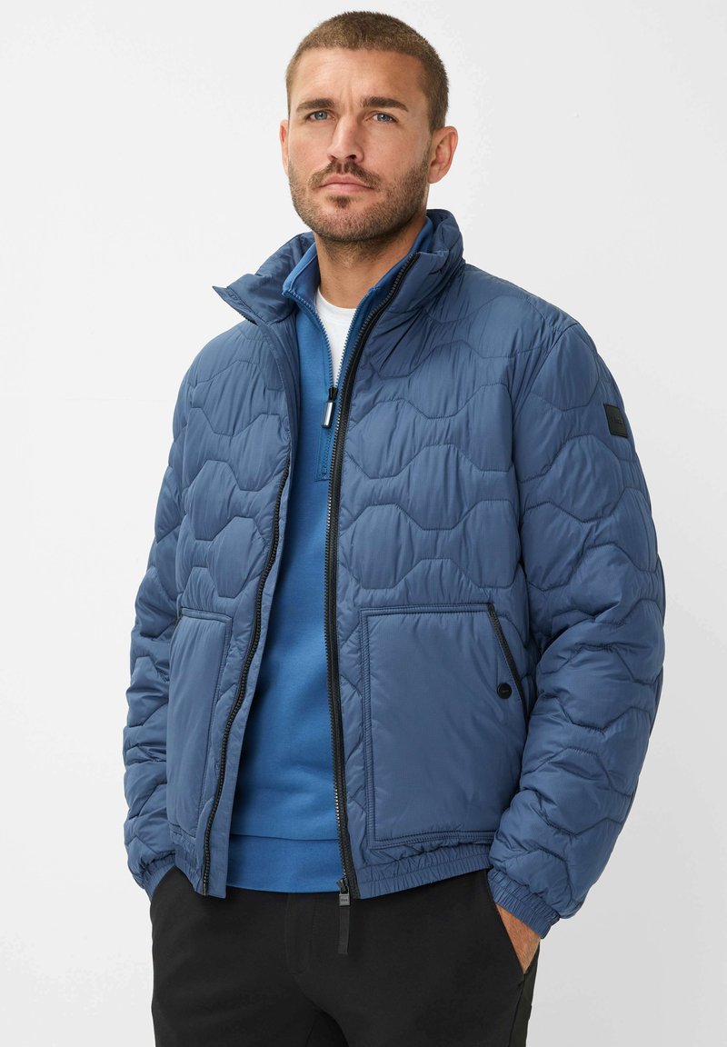 Pufferjakke i mørkeblå, quiltet mønster, letvægtsmateriale, lynlås lukning, to forlommer, høj krave, båret over en blå sweater.