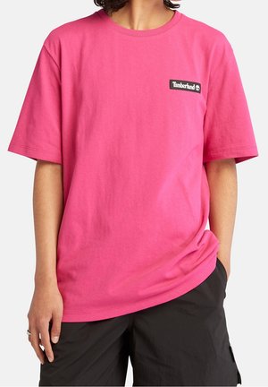 Personne portant un t-shirt Timberland rose vif à manches courtes et un short noir, une main dans la poche, sur un fond uni.