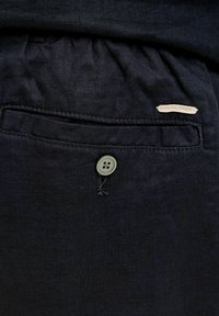 Közeli kép egy tengerészkék nadrágzsebről, amelyen gombos záródás és a zsebszél felett egy kis "Jack & Jones" címke látható.