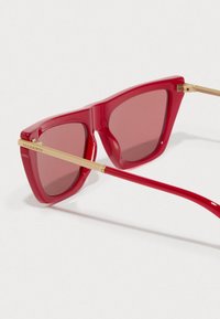 Óculos de sol em formato de gato vermelho com braços em metal dourado, incluindo o texto "ALLSAINTS" na lateral, contra um fundo branco liso.