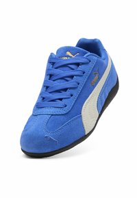 Zapatillas de ante azul con suela de goma blanca y negra; cuenta con cordones redondos y un acento de logo dorado en el lateral.