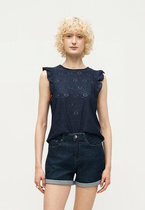 Persona bionda con capelli ricci che indossa una canotta blu navy con occhielli e spalle arricciate e pantaloncini di jeans scuri risvoltati.