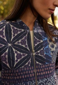 Chaqueta bomber acolchada en azul marino con intrincados patrones geométricos en púrpura y azul, con cremallera frontal y cuello alzado.